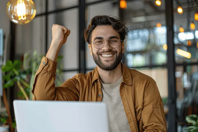 A young man raises his fist behind a laptop while celebrating in a modern office.