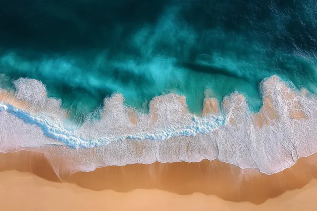 Aerial view of turquoise ocean waves breaking over golden sand, creating foam patterns in a serene summer coastal scene.