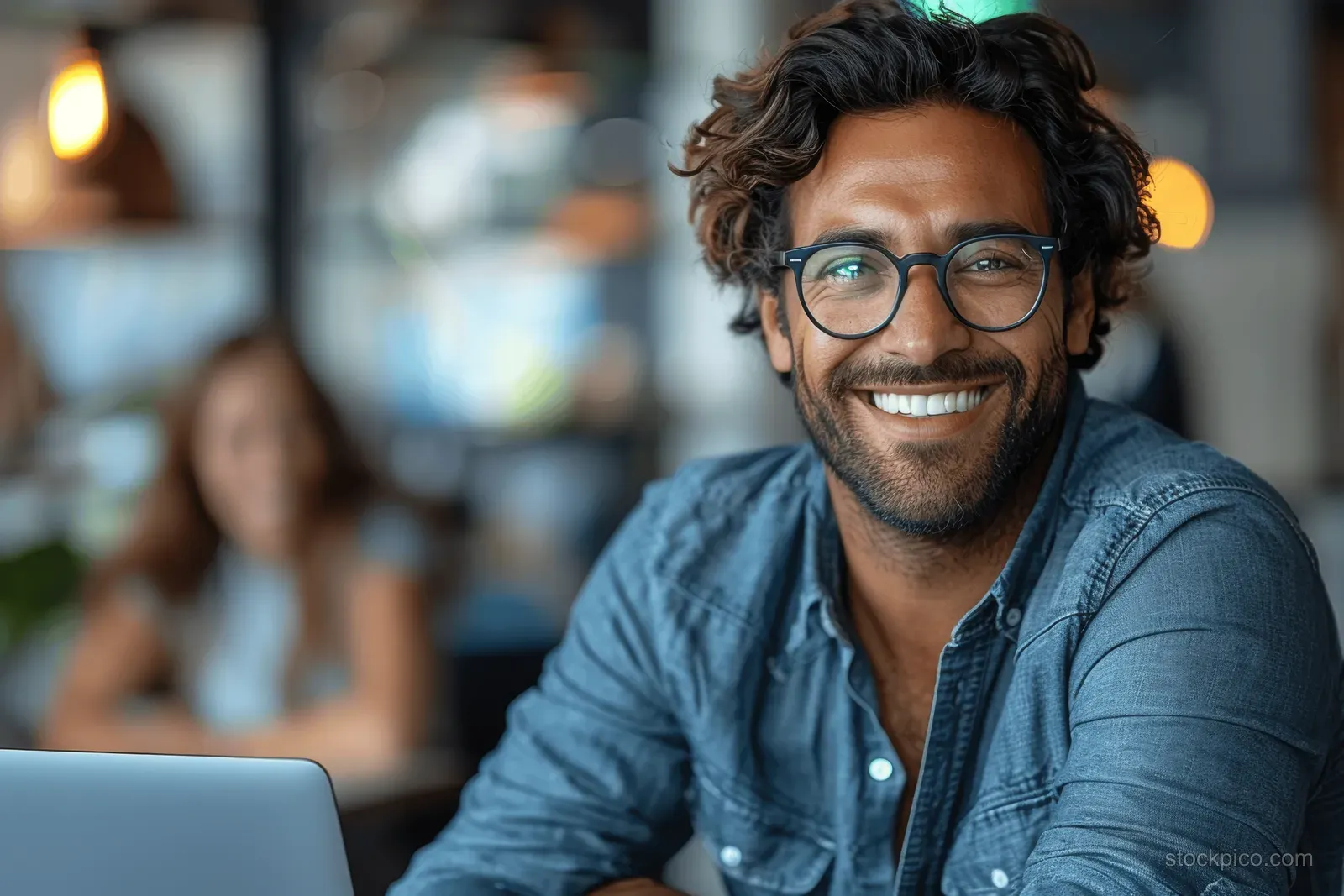 A smiling man in a denim shirt sits at an office table using a laptop during a meeting with blurred coworkers behind.