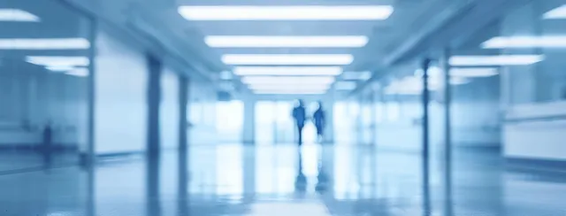 Blurred blue hospital corridor with bright ceiling lights and distant doorway visible through soft focus.