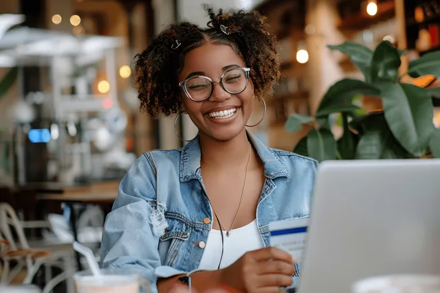 A joyful Black woman makes a secure online payment using a laptop and smartphone in a cozy cafe setting.
