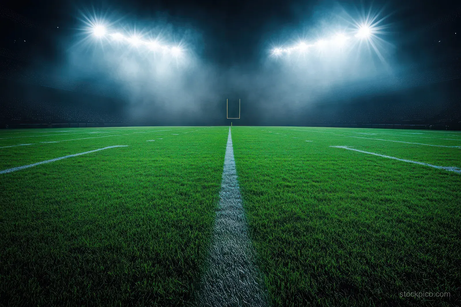 Night football field with floodlights shining on vivid green grass and a centered yard line fading into darkness.
