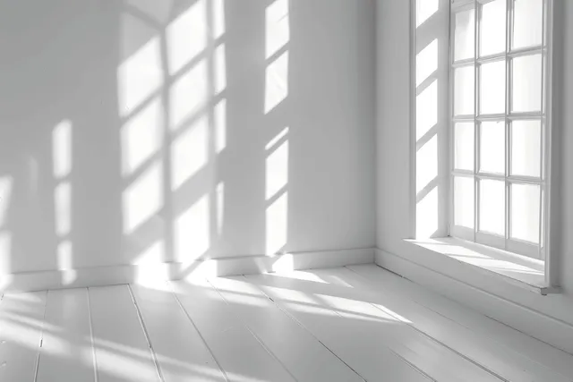 Sunlight streams through tall windows, casting abstract shadows across an empty white room interior.