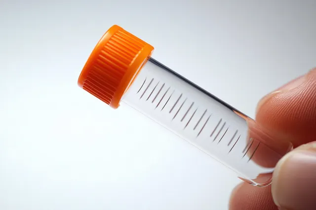 A hand holds a clear plastic test tube with an orange cap showing measurement markings on a white background.