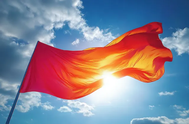A red Labour Party flag billows in the wind under a dramatic blue sky with sunlit highlights and textured fabric motion.