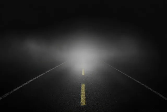 An empty dark asphalt road at night disappears into thick fog, marked by a fading yellow dashed center line.