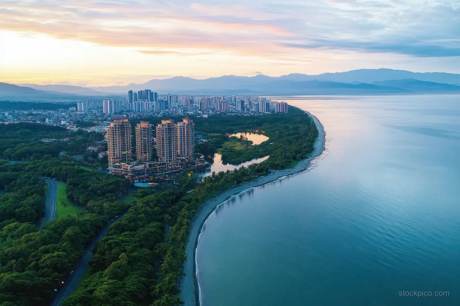 Aerial view of tall buildings beside a green shoreline and calm sea at sunset.