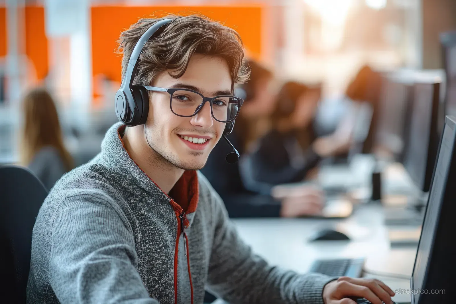 Young man in glasses and headset typing on a desktop computer at a busy call-center desk.