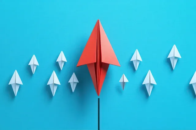 Red paper airplane leads white flock forward on vivid blue background