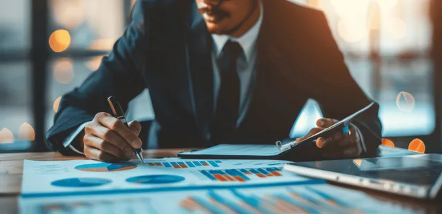 Businessman writing with a pen over financial documents while reviewing colorful chart graphs on an office desk.