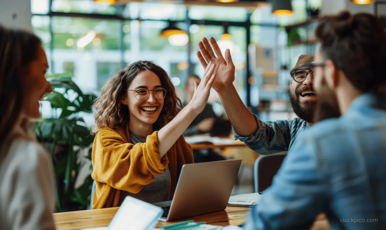 Smiling coworkers high-five across a conference table during a lively office meeting with laptops and papers.