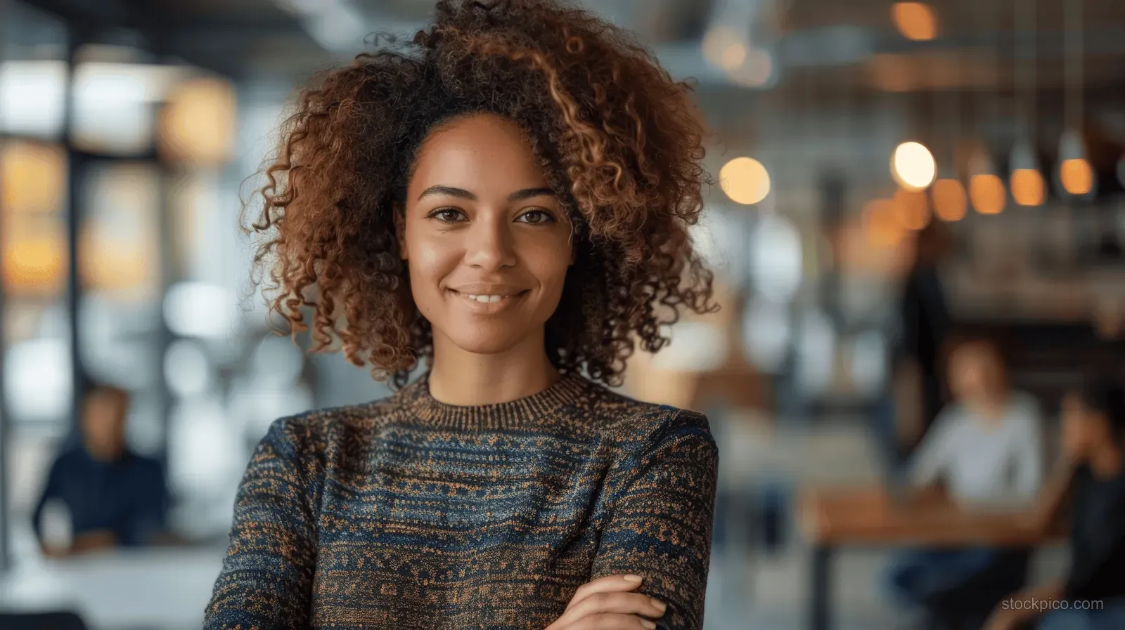 Smiling businesswoman with curly hair stands in an office with colleagues working behind her in soft focus.