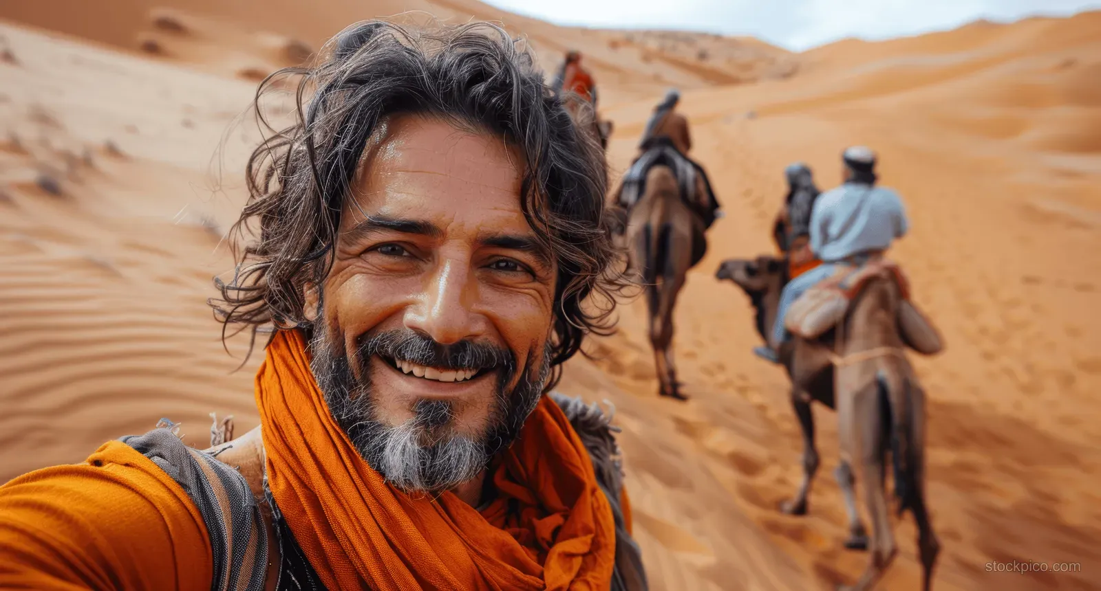 Smiling man in an orange scarf takes a selfie on a camel ride in the desert.