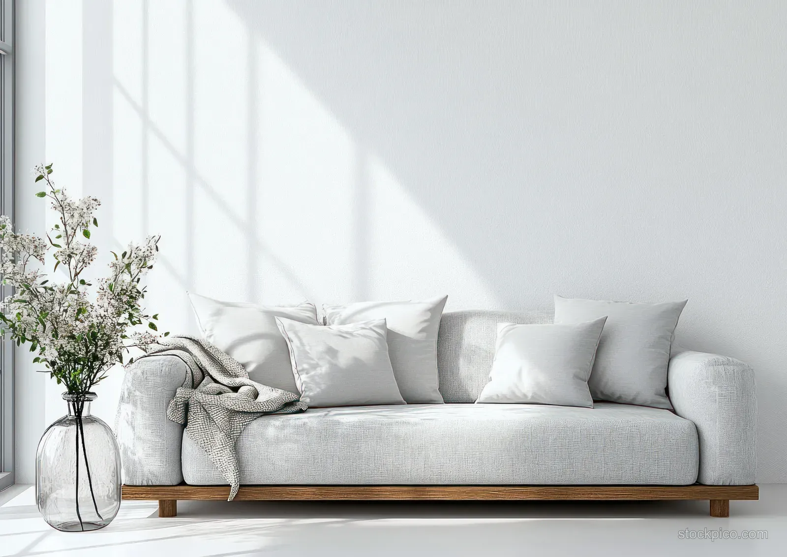 Minimalist gray sofa with white pillows and fresh flowers in a tall vase beside a bright large window.