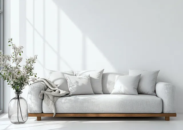 Minimalist gray sofa with white pillows and fresh flowers in a tall vase beside a bright large window.