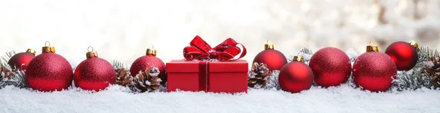 Red Christmas ornaments and a wrapped gift box are arranged on snowy ground with blurred holiday lights in the background.