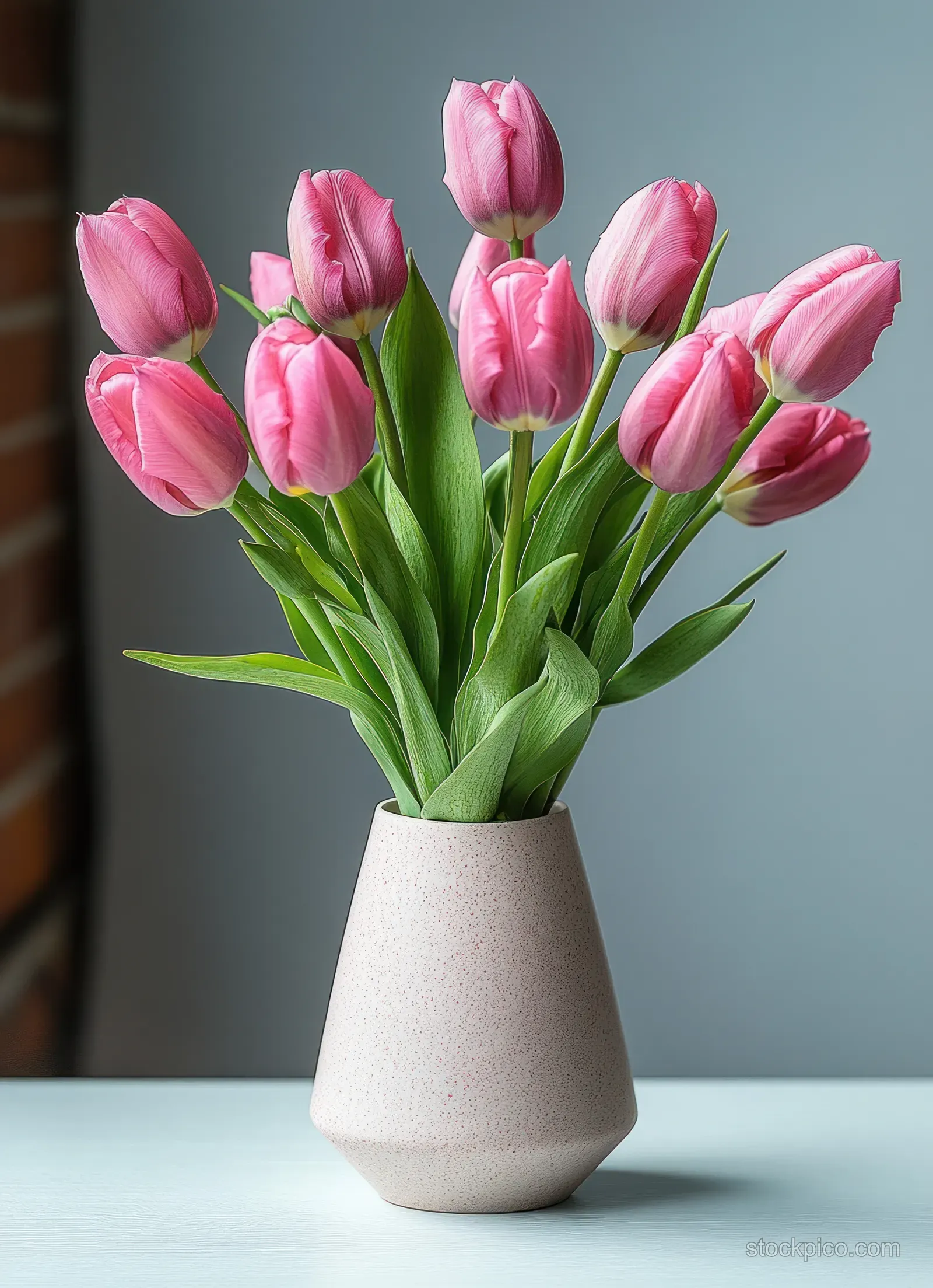 Pink tulip bouquet with green leaves in a light pink vase on a white surface against a neutral gray background.