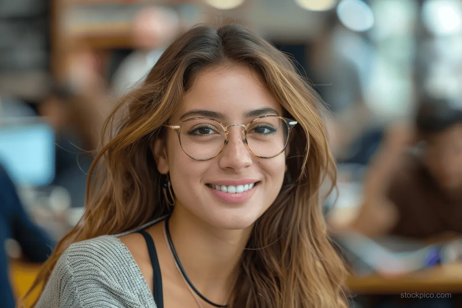 Smiling young woman wearing glasses sits in a busy office.