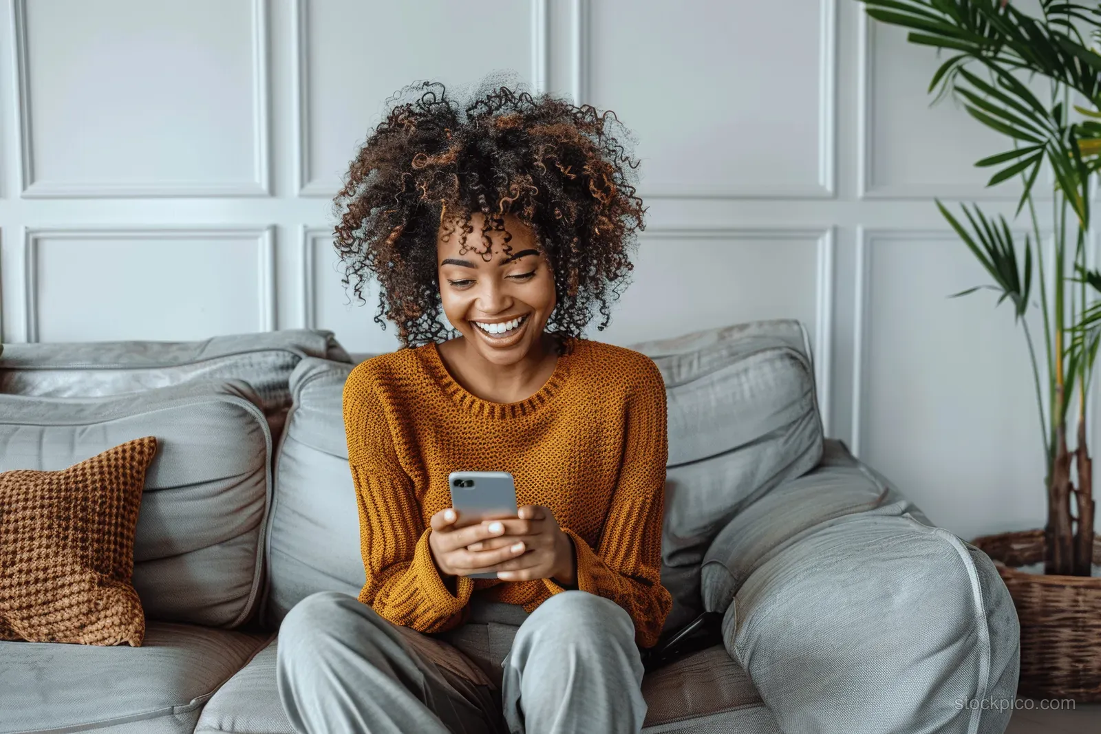 Young Black woman sitting on a gray sofa indoors, holding a smartphone and smiling surprised.