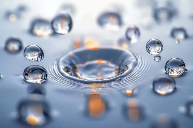Clear water droplets hover above a ripple pool with glowing orange light reflections on a glossy surface.