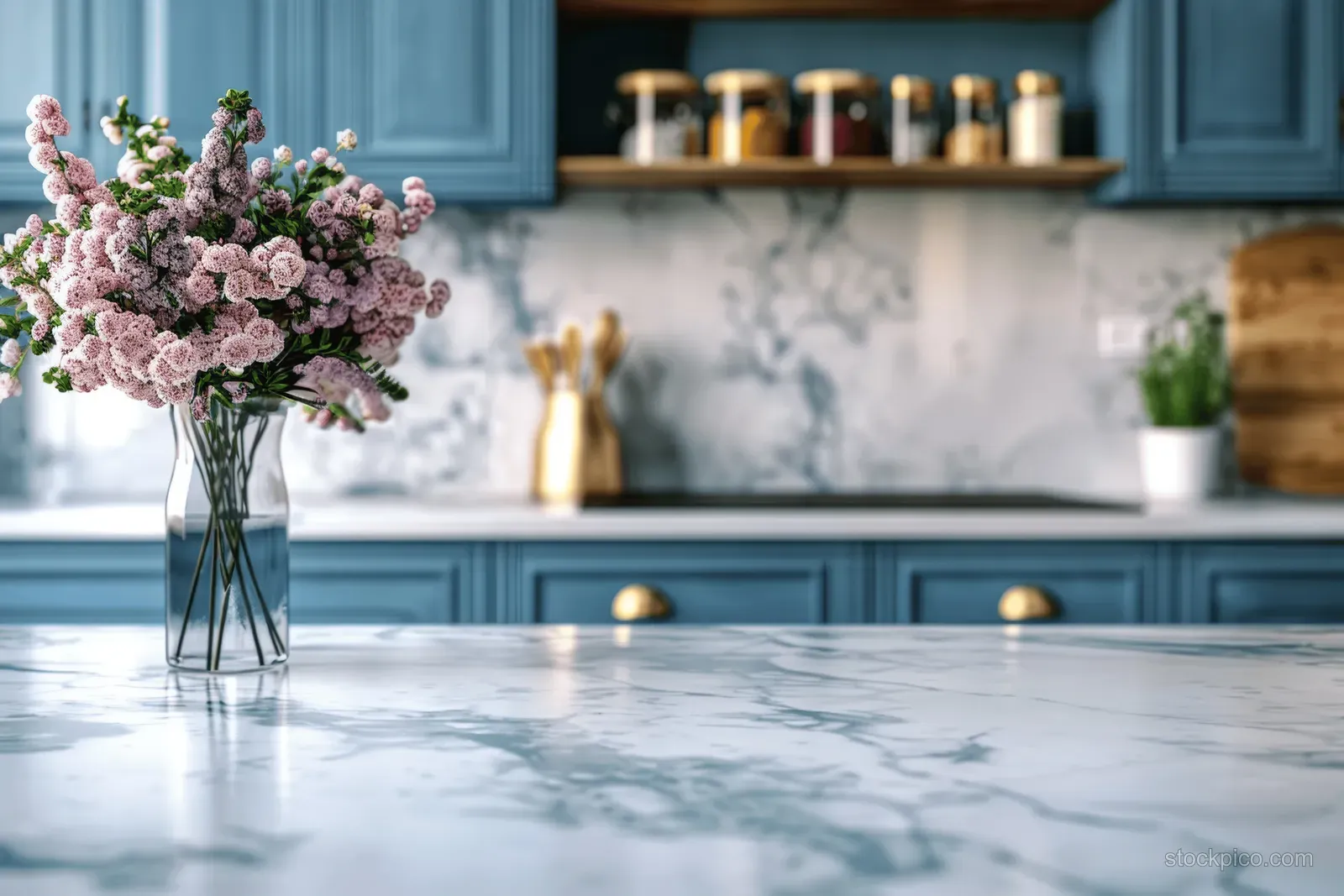 White marble countertop with a glass vase of pink flowers, set before blurred blue kitchen cabinets and a marble backsplash.