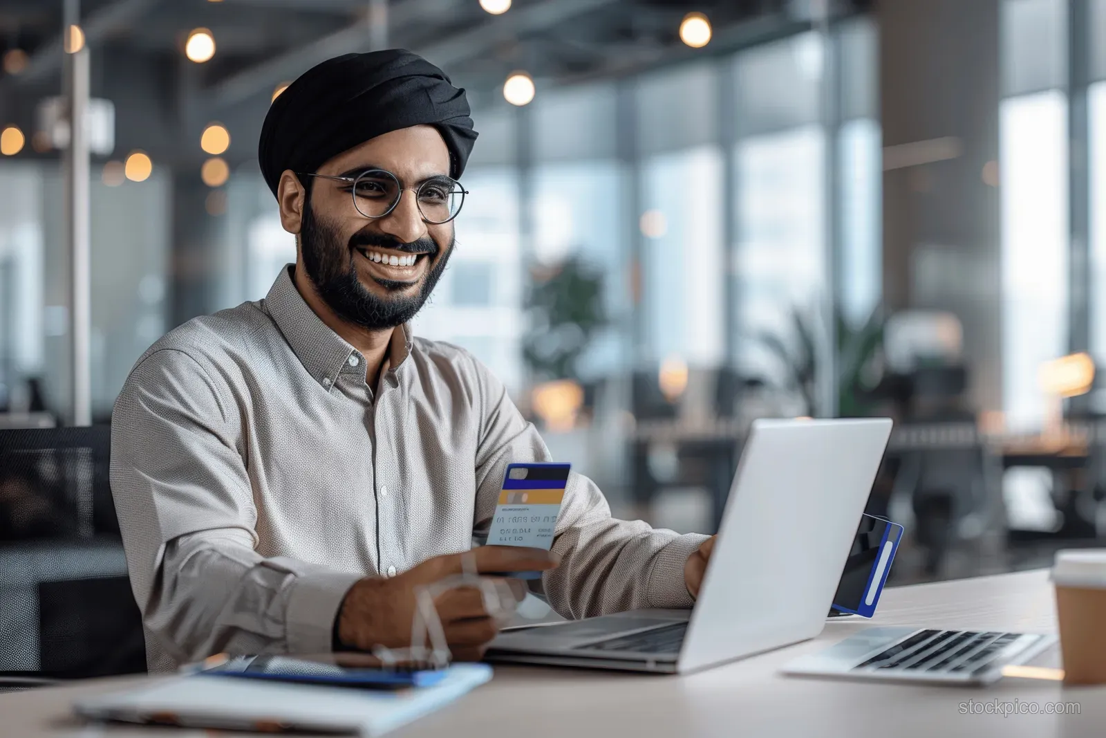 Young businessman at a modern office desk holds a credit card and looks at a smartphone near a laptop.