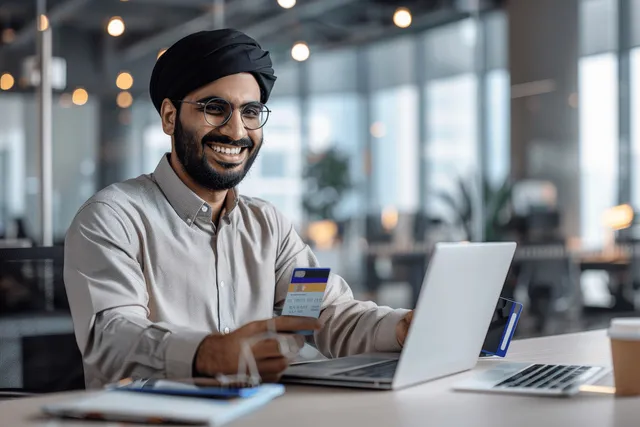 Young businessman at a modern office desk holds a credit card and looks at a smartphone near a laptop.