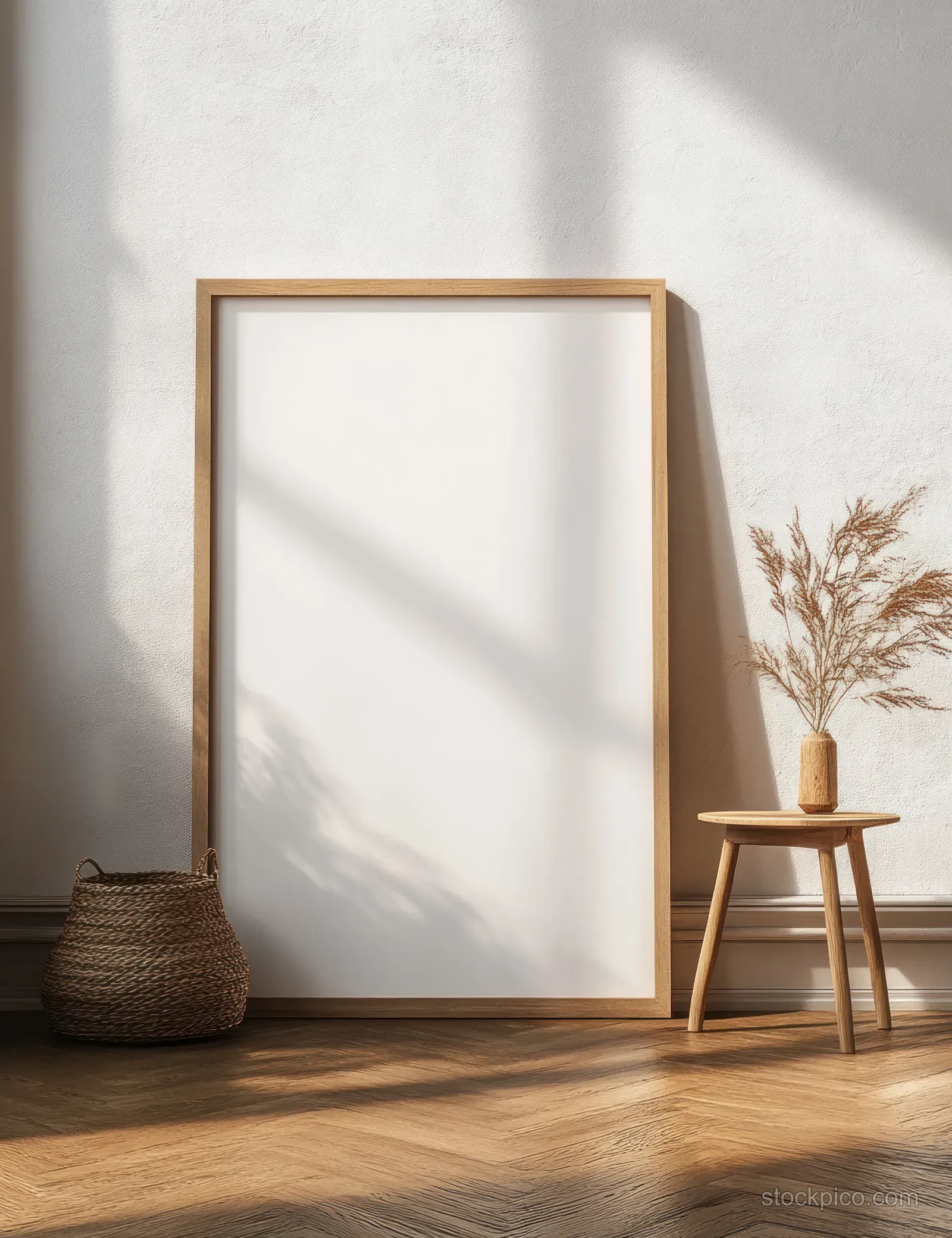 Blank canvas in a minimal wooden frame leans indoors with sunlight shadows, a stool, and dried grass nearby.
