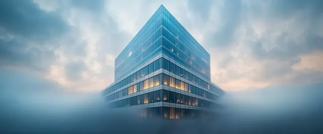 A glass office tower rises through thick morning fog with warm interior lights under cloudy skies.