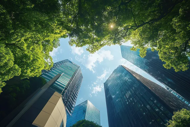 Upward view of modern glass skyscrapers surrounded by leafy trees beneath a blue sky.
