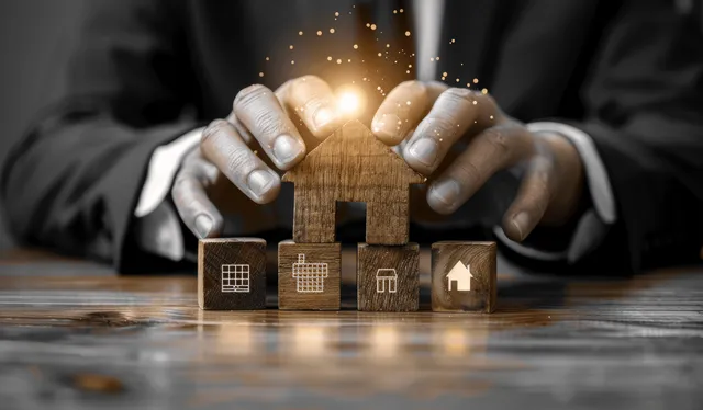 Hands shield miniature houses on a table with a glowing security light effect.