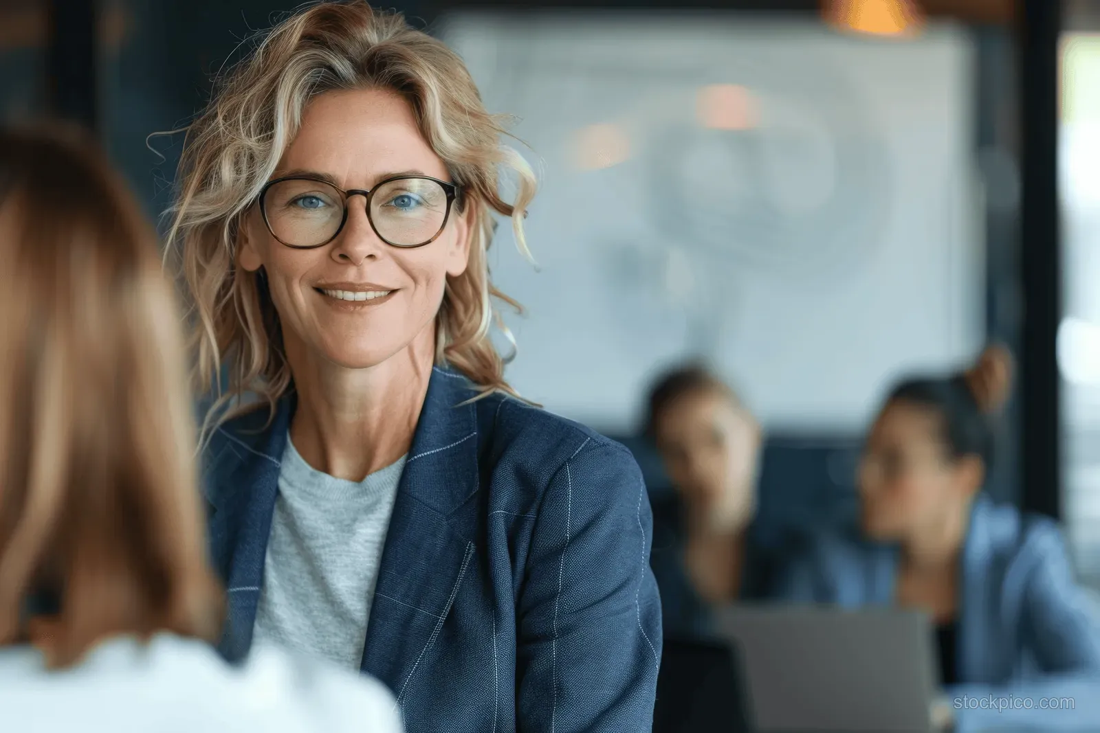 Smiling businesswoman in a blue blazer presenting to coworkers during an office meeting with blurred background.