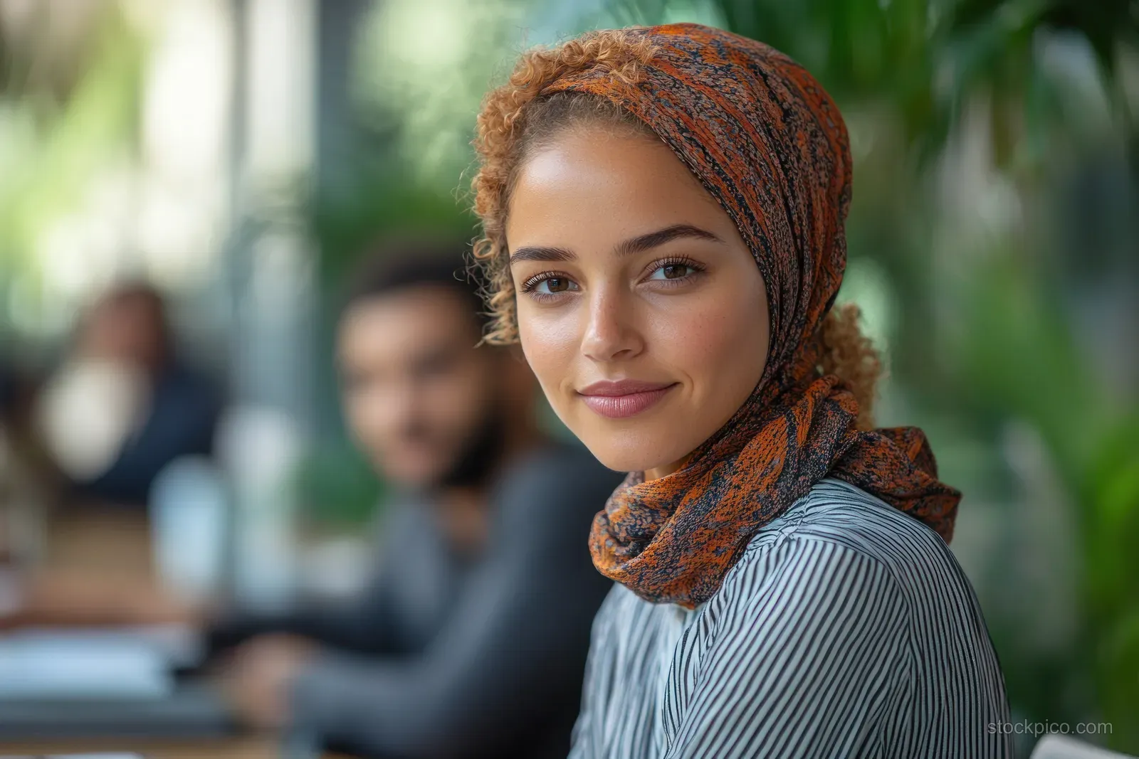 Indian woman in a patterned headscarf stands in a modern office during a team meeting while others sit blurred behind