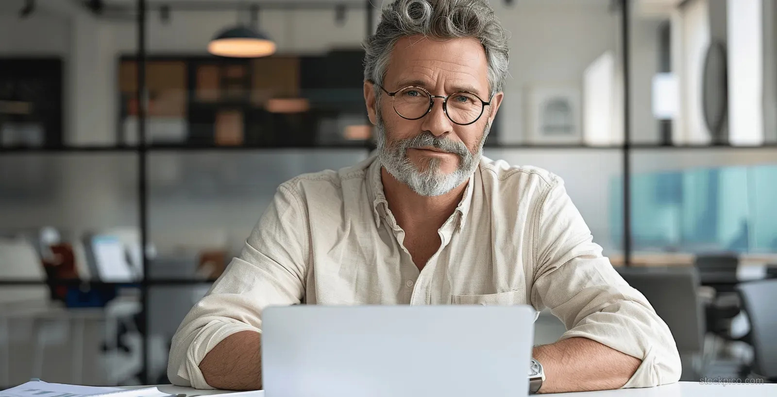 A middle-aged man with glasses works on a laptop at a desk in a glass-walled office.