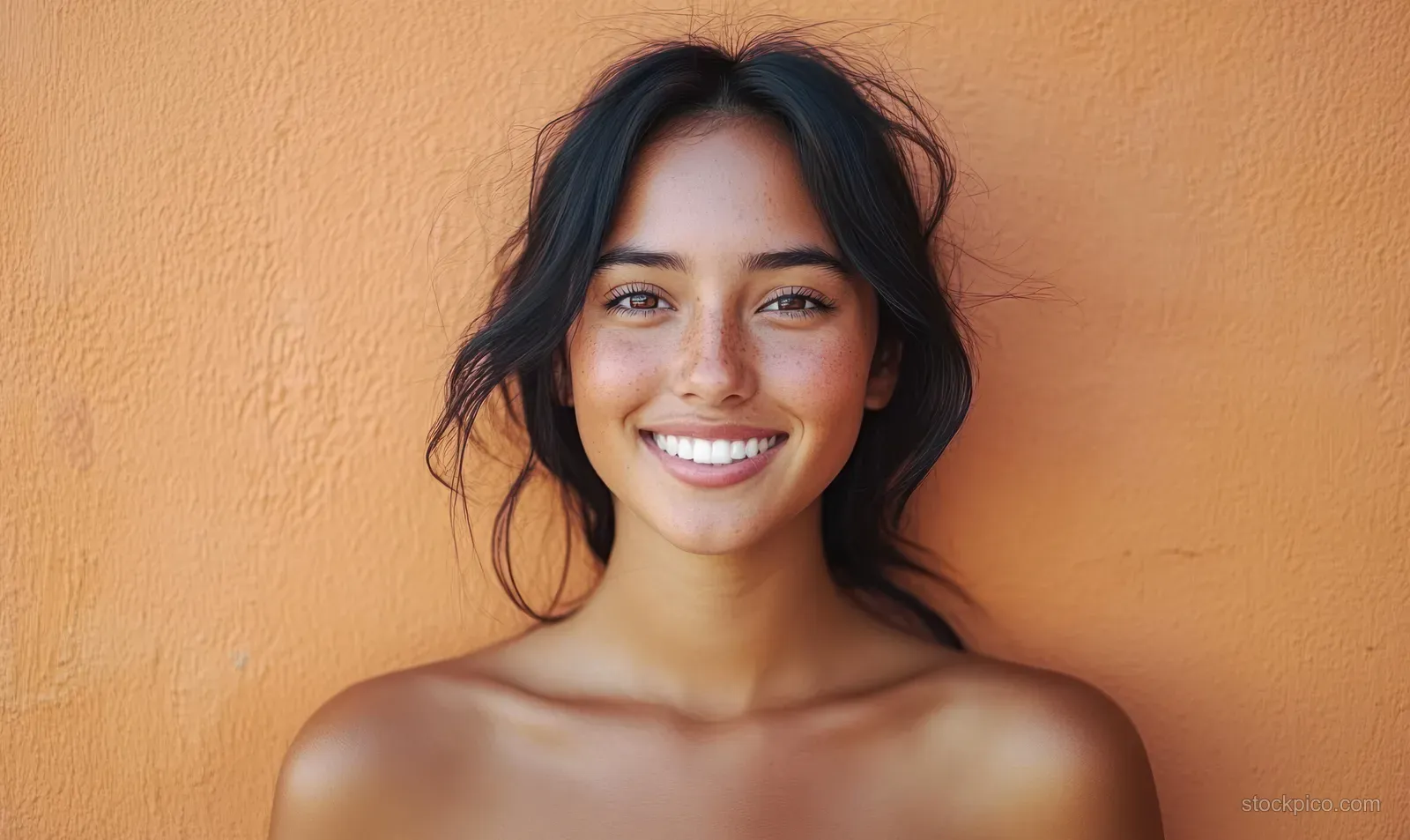Young woman with dark hair smiles at the camera against a textured orange wall.