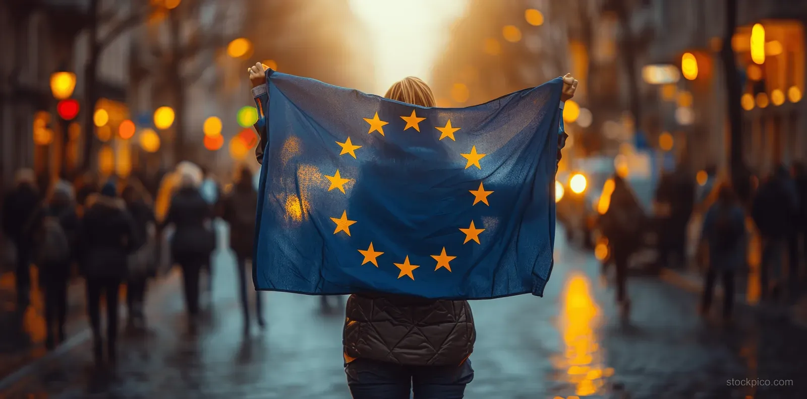 Young woman holding a European Union flag on a rainy city street at sunset with blurred crowd lights behind.