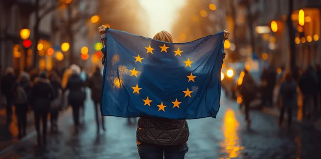 Young woman holding a European Union flag on a rainy city street at sunset with blurred crowd lights behind.