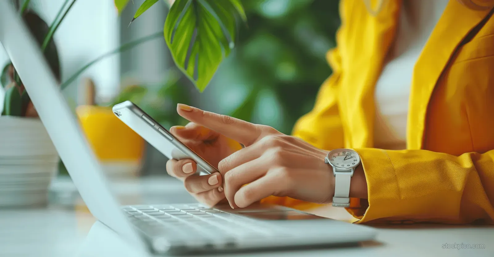 Woman in a yellow blazer holds a smartphone while typing on a laptop keyboard at an office desk.