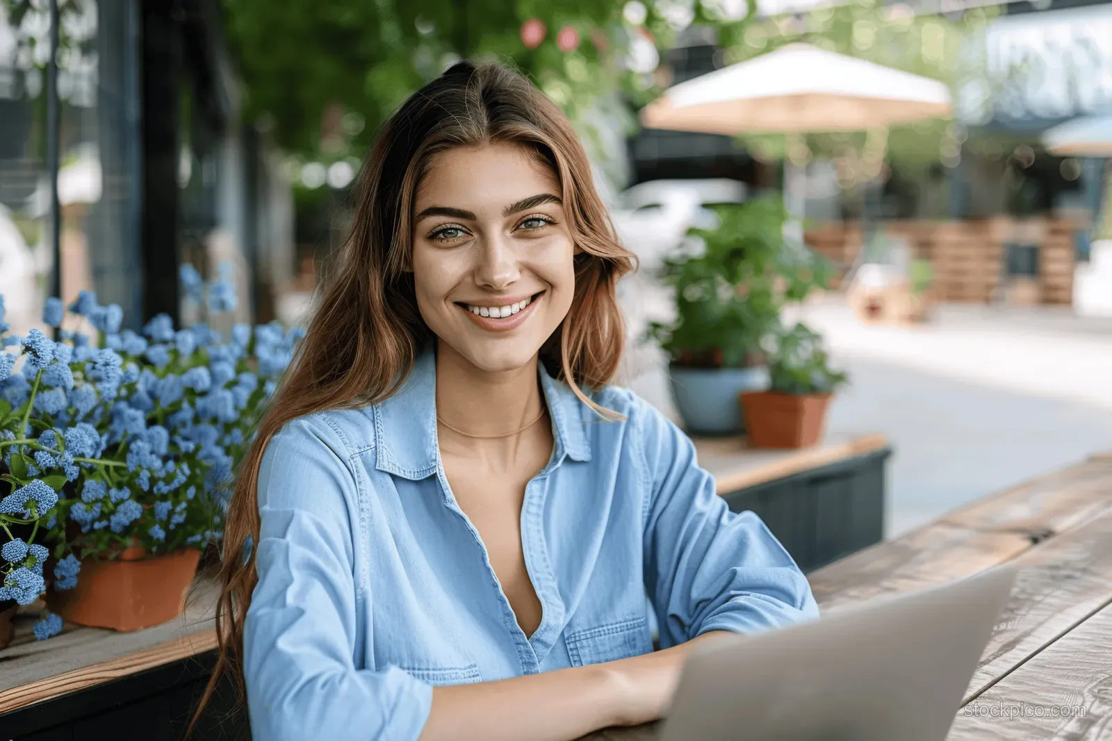 Smiling young businesswoman sits outdoors with a laptop, video calling beside glass windows and flower planters.