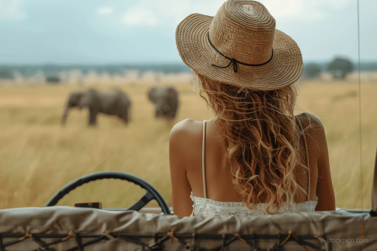 A woman in a straw hat sits in an open safari jeep looking toward grazing elephants across the savanna.