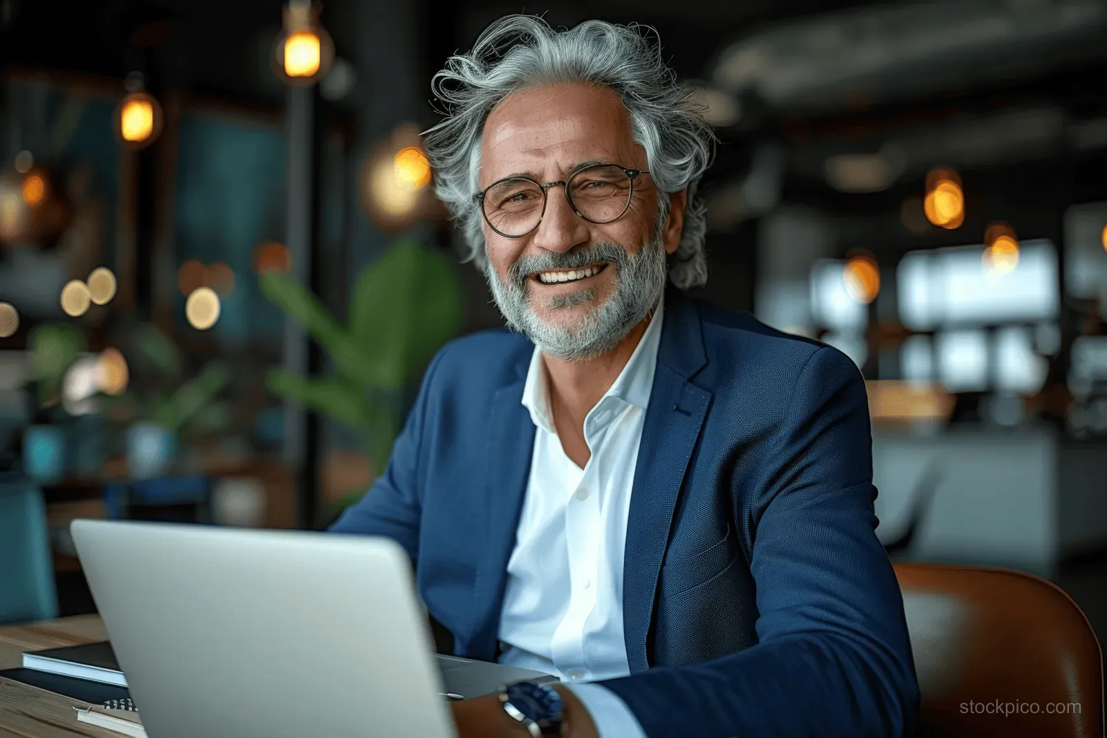 Gray-haired man in blue suit smiles at a desk while typing on a laptop in a modern office.