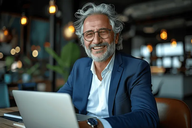 Gray-haired man in blue suit smiles at a desk while typing on a laptop in a modern office.