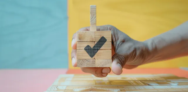 A hand holds a wooden block printed with a checkmark against a colorful background.