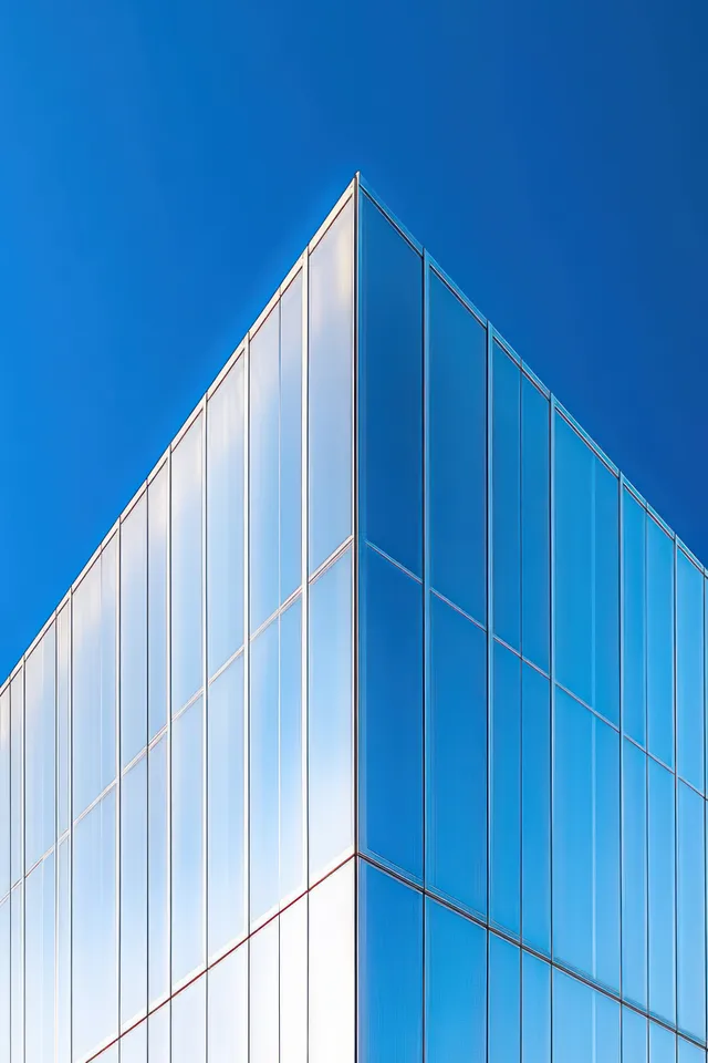 The corner of a modern building shows reflective glass panels rising vertically under a clear blue sky.
