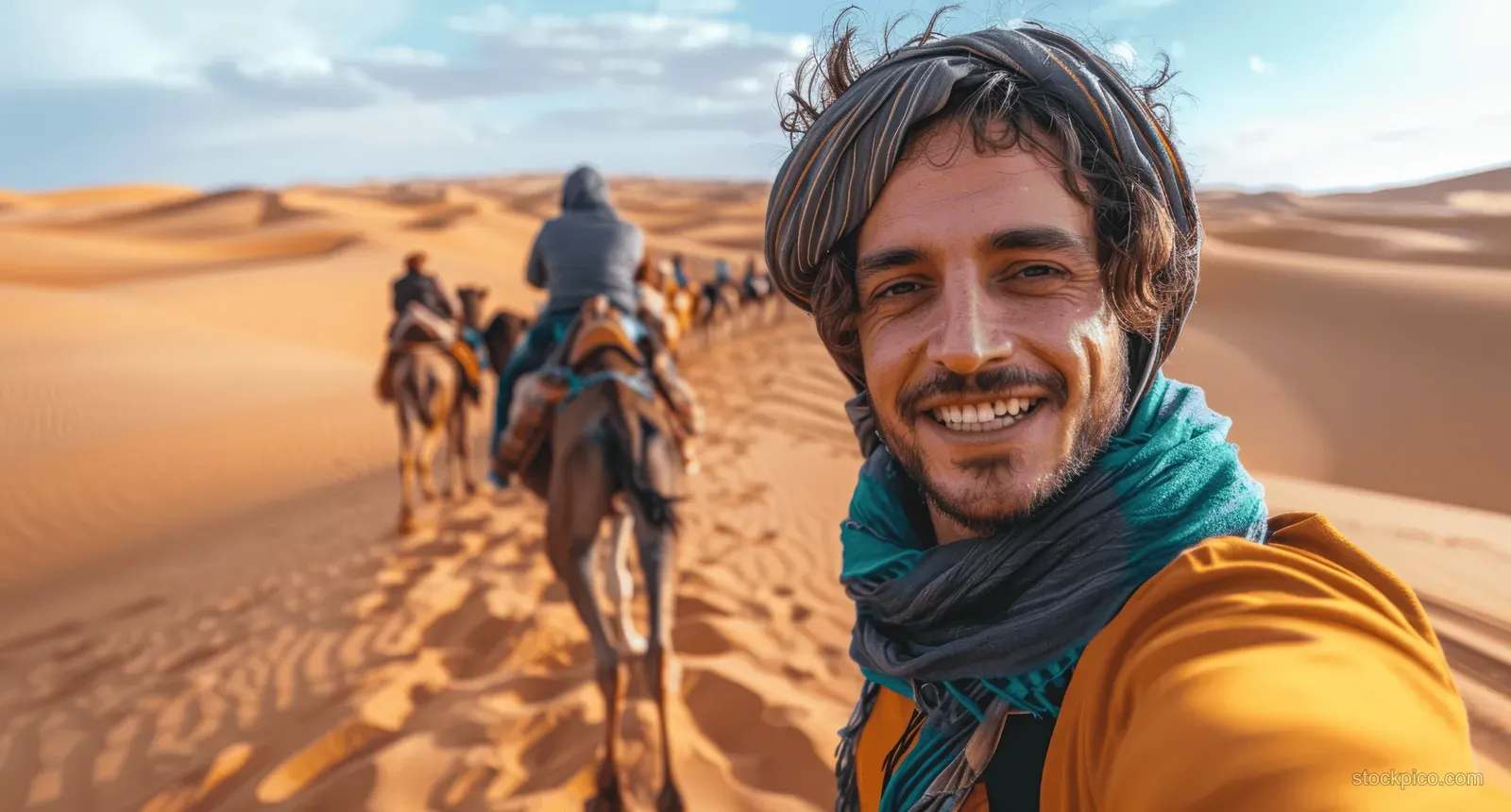 A man on a camel takes a selfie in a desert with other riders visible in the background.