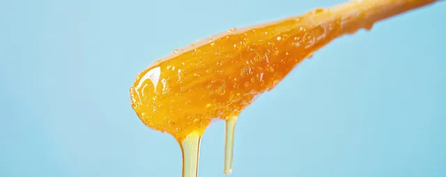 Golden wax drips from a wooden stick on blue backdrop for hair removal