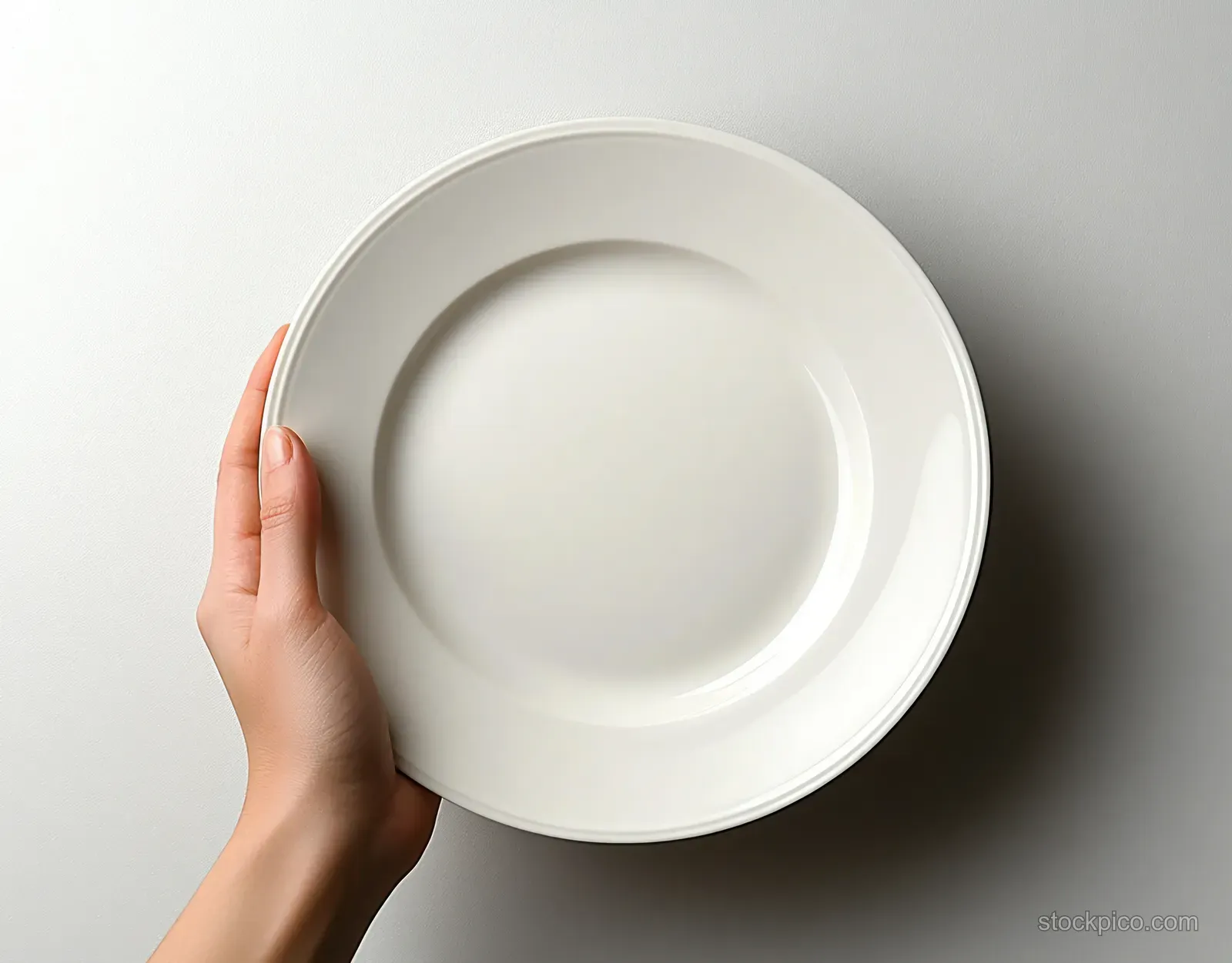 A hand holds an empty white round ceramic plate against a plain white background.