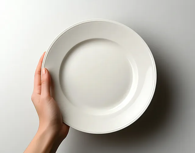 A hand holds an empty white round ceramic plate against a plain white background.