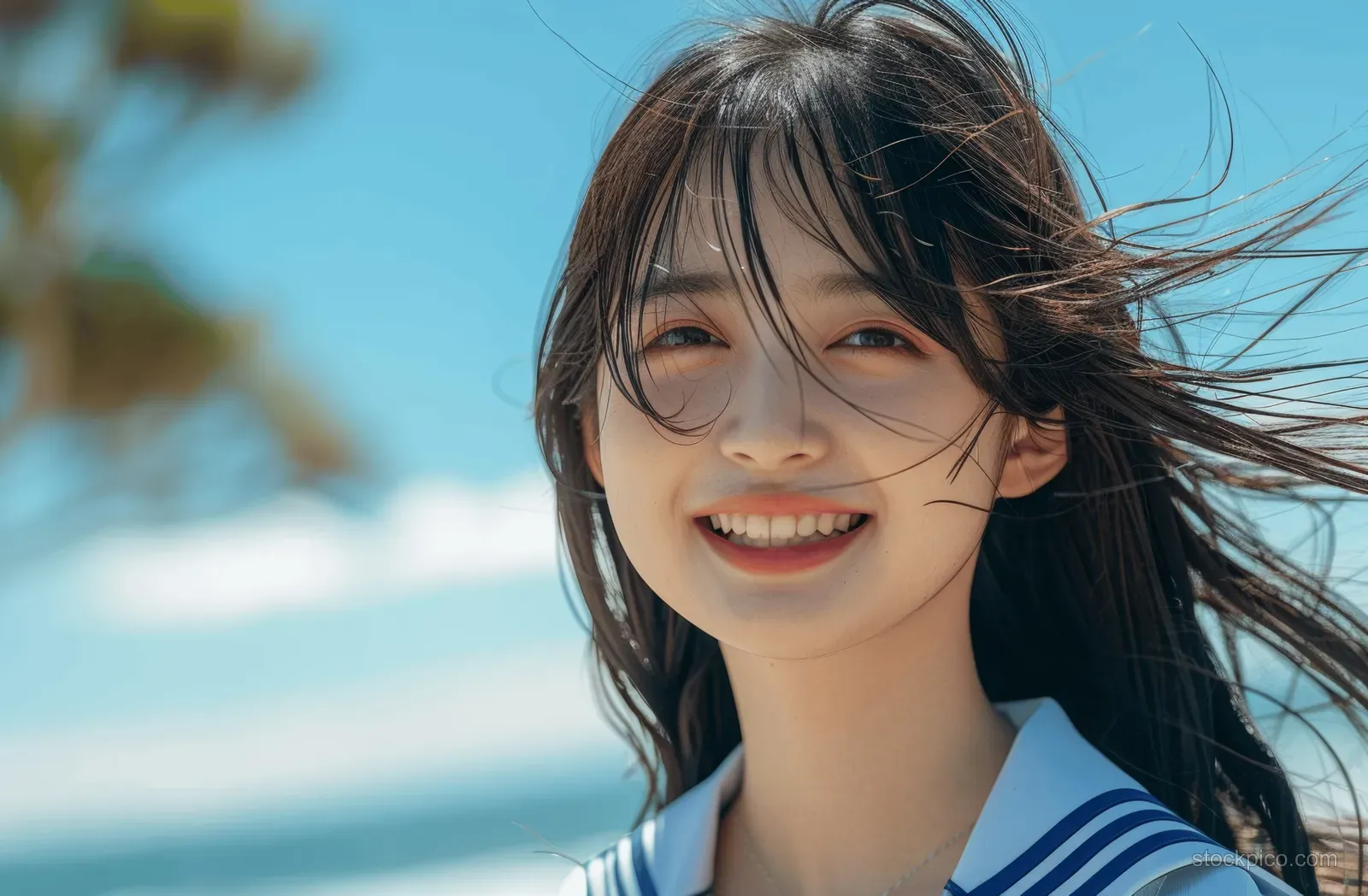 Smiling Japanese schoolgirl in a blue sailor uniform outdoors with wind-tossed hair under a clear summer sky.