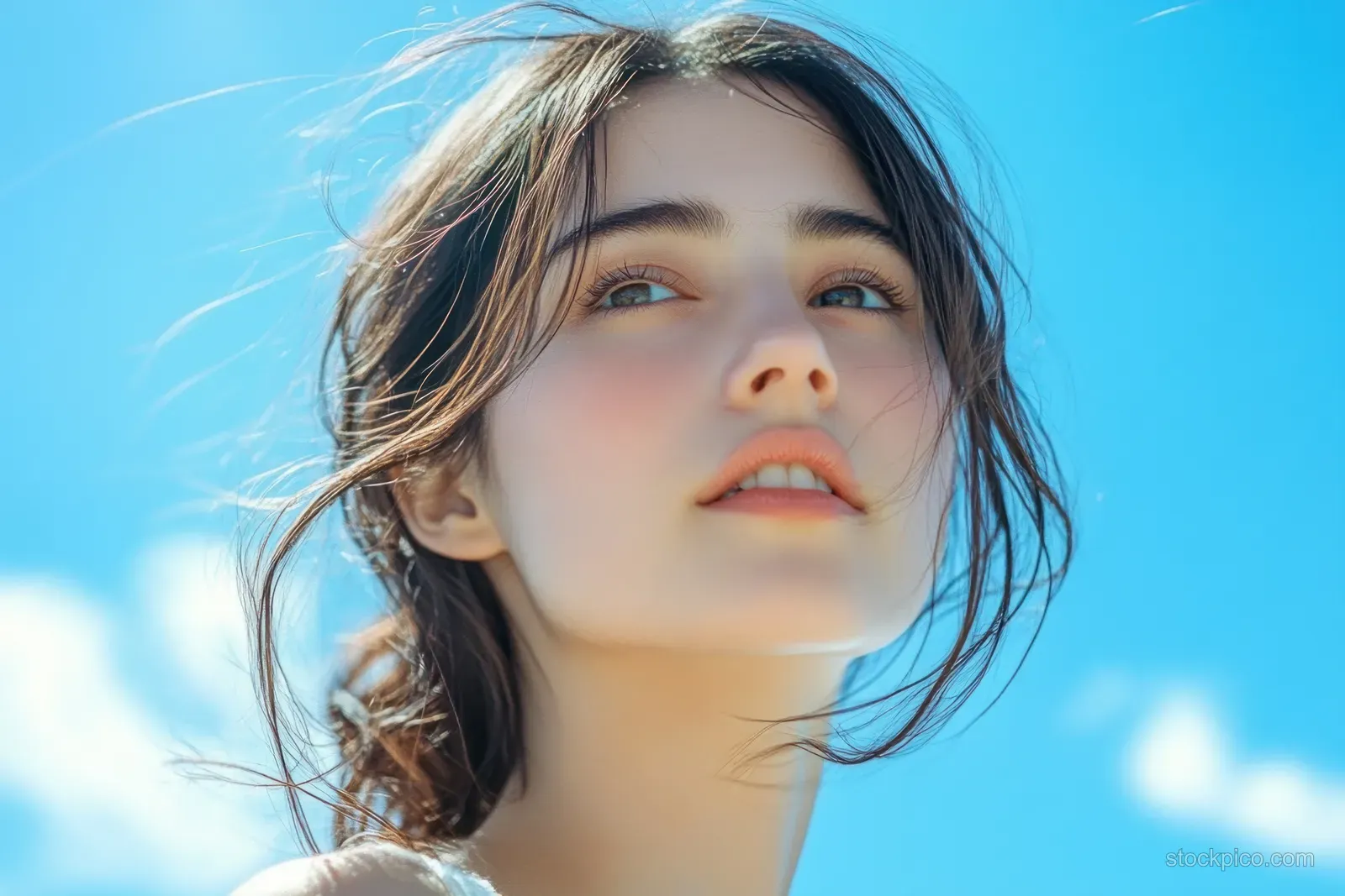 Young woman looks up smiling outdoors as wind lifts her hair against a bright blue sky.